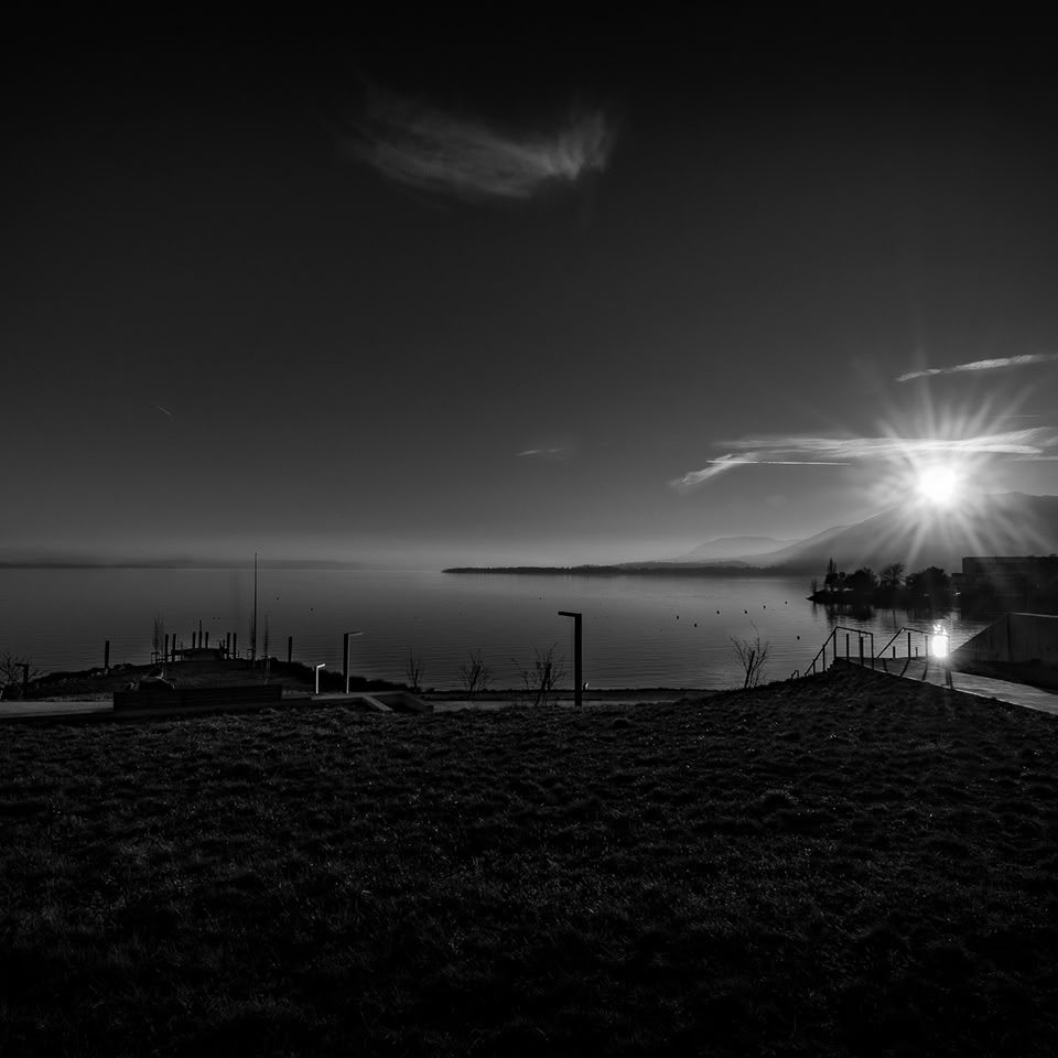 Paysage en noir et blanc avec un lac, un ponton, des montagnes à l'horizon et le soleil se levant ou se couchant.