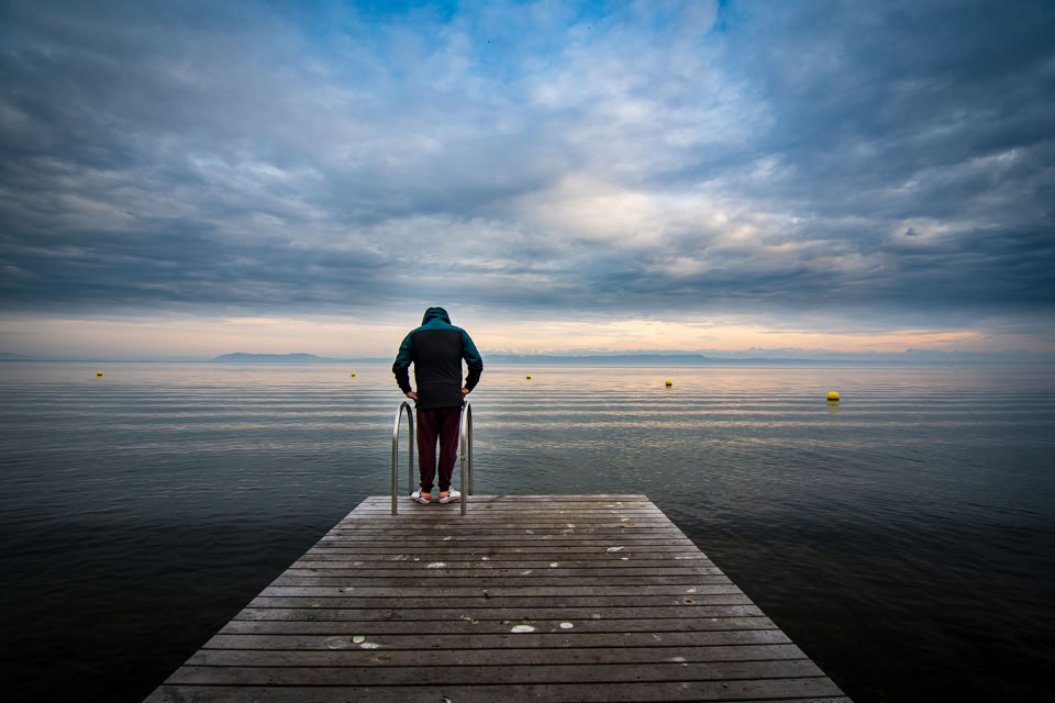 Person standing on a wooden dock facing a calm body of water under a cloudy sky.