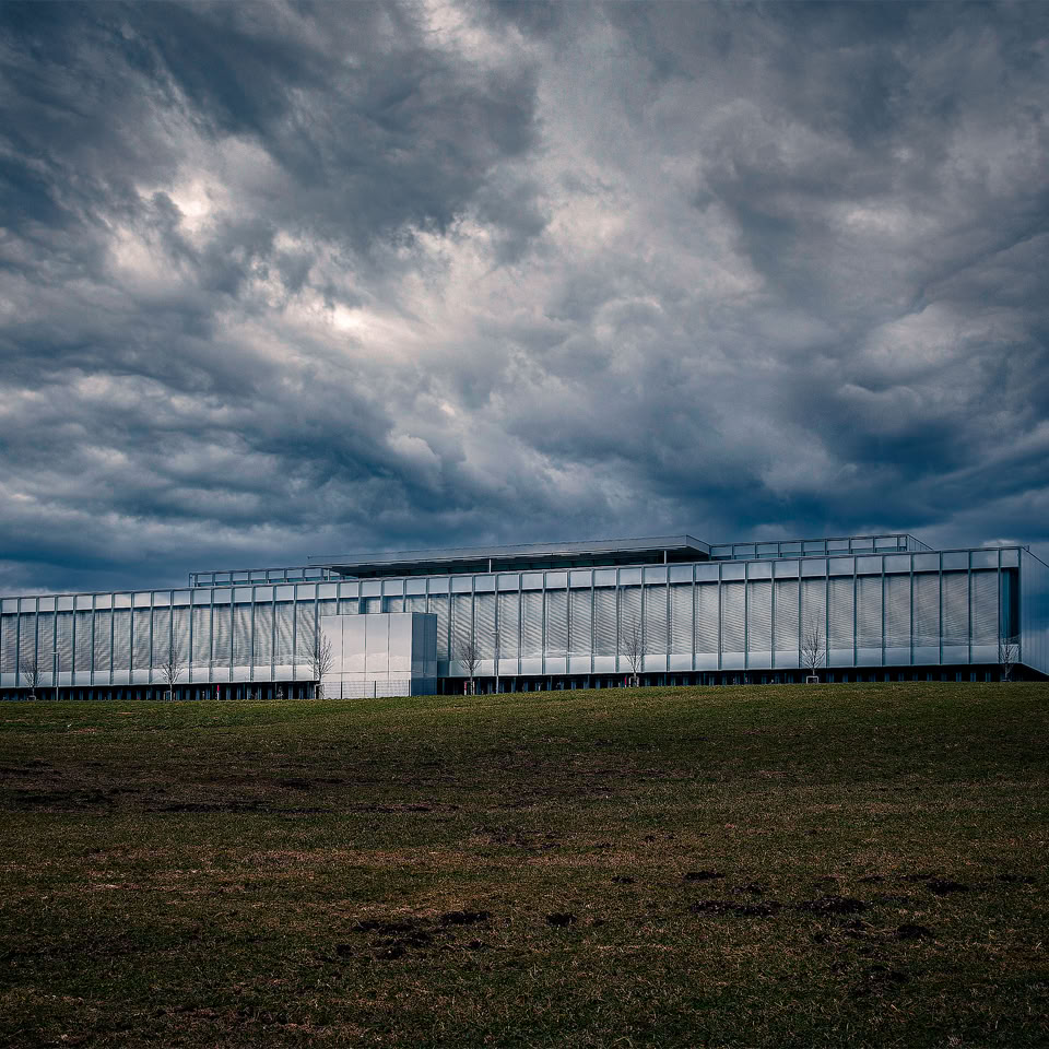 Bâtiment moderne en verre sur une colline sous un ciel nuageux et sombre.