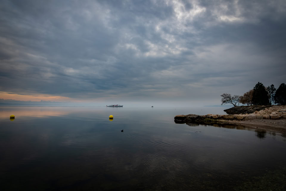 A calm lake with a cloudy sky, distant yellow buoys, and a rocky shoreline with trees on the right.