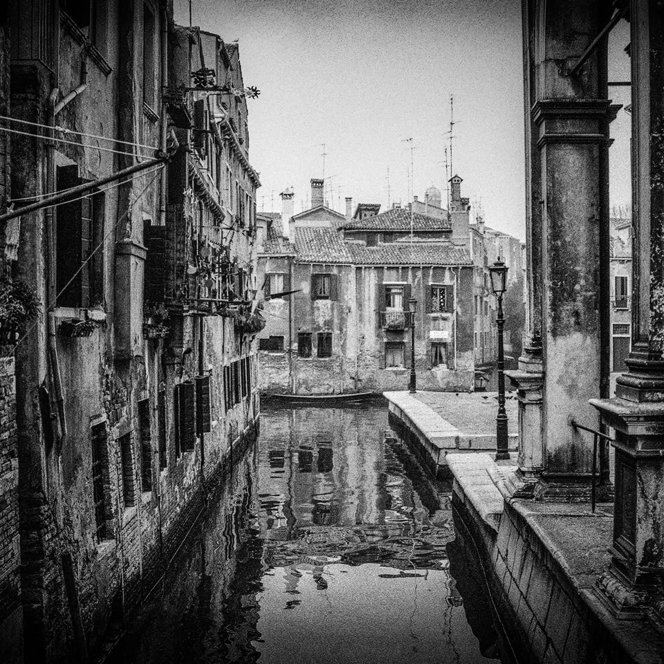 Vue en noir et blanc d'un canal étroit bordé de bâtiments anciens avec des détails architecturaux, se terminant par une place avec des structures historiques en arrière-plan.