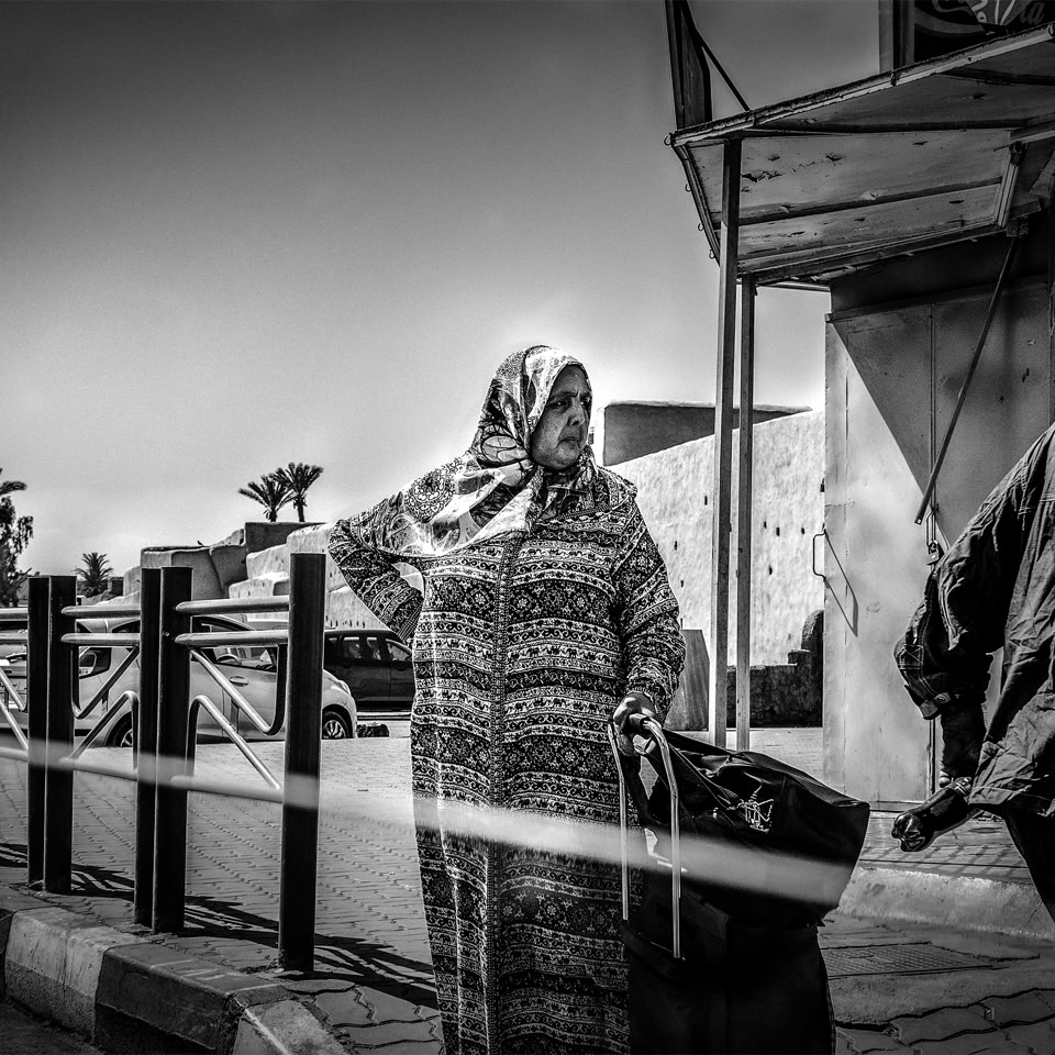 Une femme en robe traditionnelle marche sur un trottoir près d'une barrière, avec un homme de dos à droite et un bâtiment en arrière-plan. L'image est en noir et blanc.
