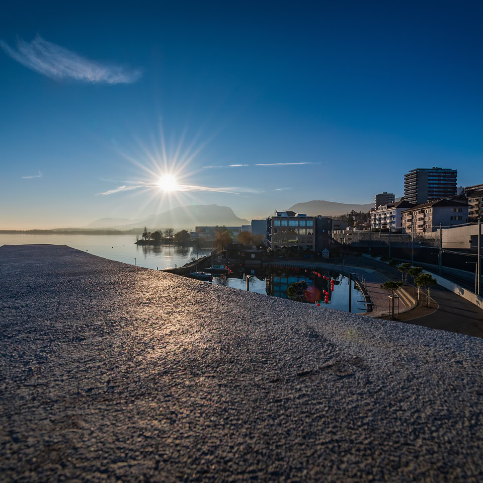 Vue d'un quai au bord de l'eau avec le soleil se levant à l'horizon, des bâtiments en arrière-plan et un ciel dégagé.