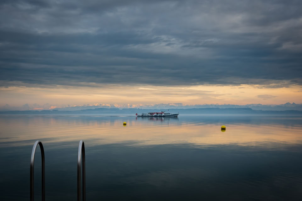 Vue d'un lac avec un ciel nuageux, un bateau à l'horizon, et des bouées jaunes flottant sur l'eau.
