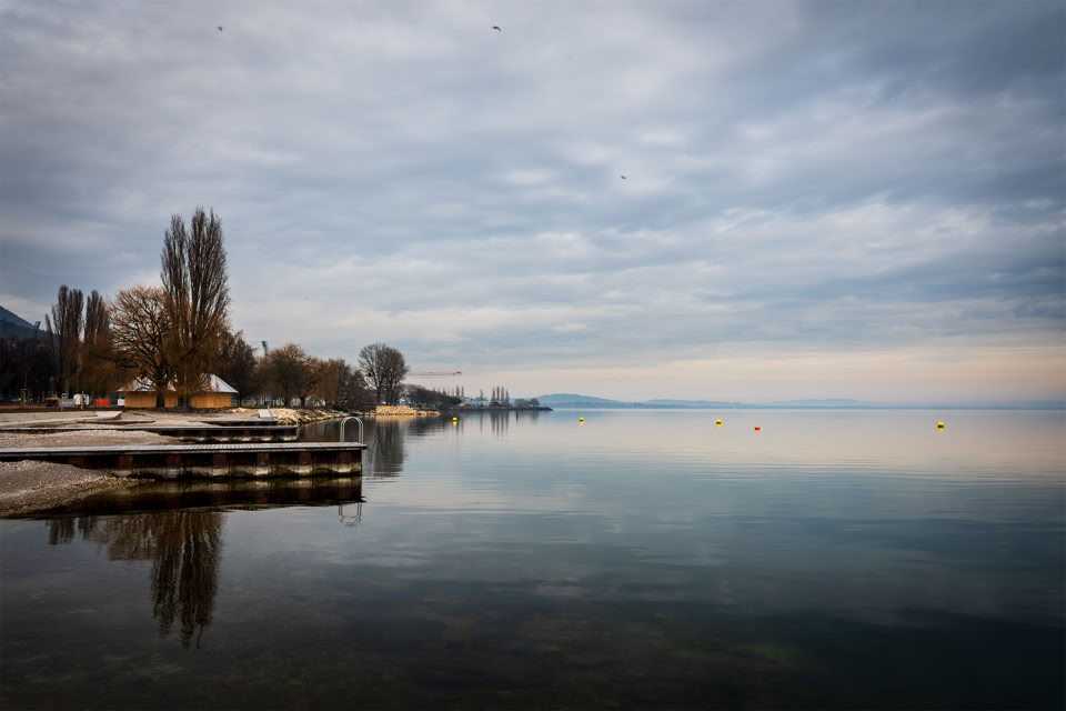 Paysage lacustre avec un ciel nuageux, des arbres dénudés sur la rive gauche et un quai en bois.